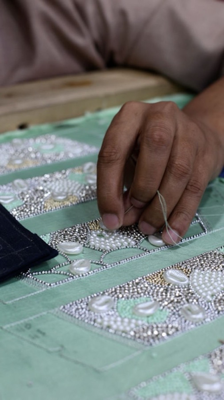An artisan's hand stitching beadwork onto fabric stretched flat on a workbench