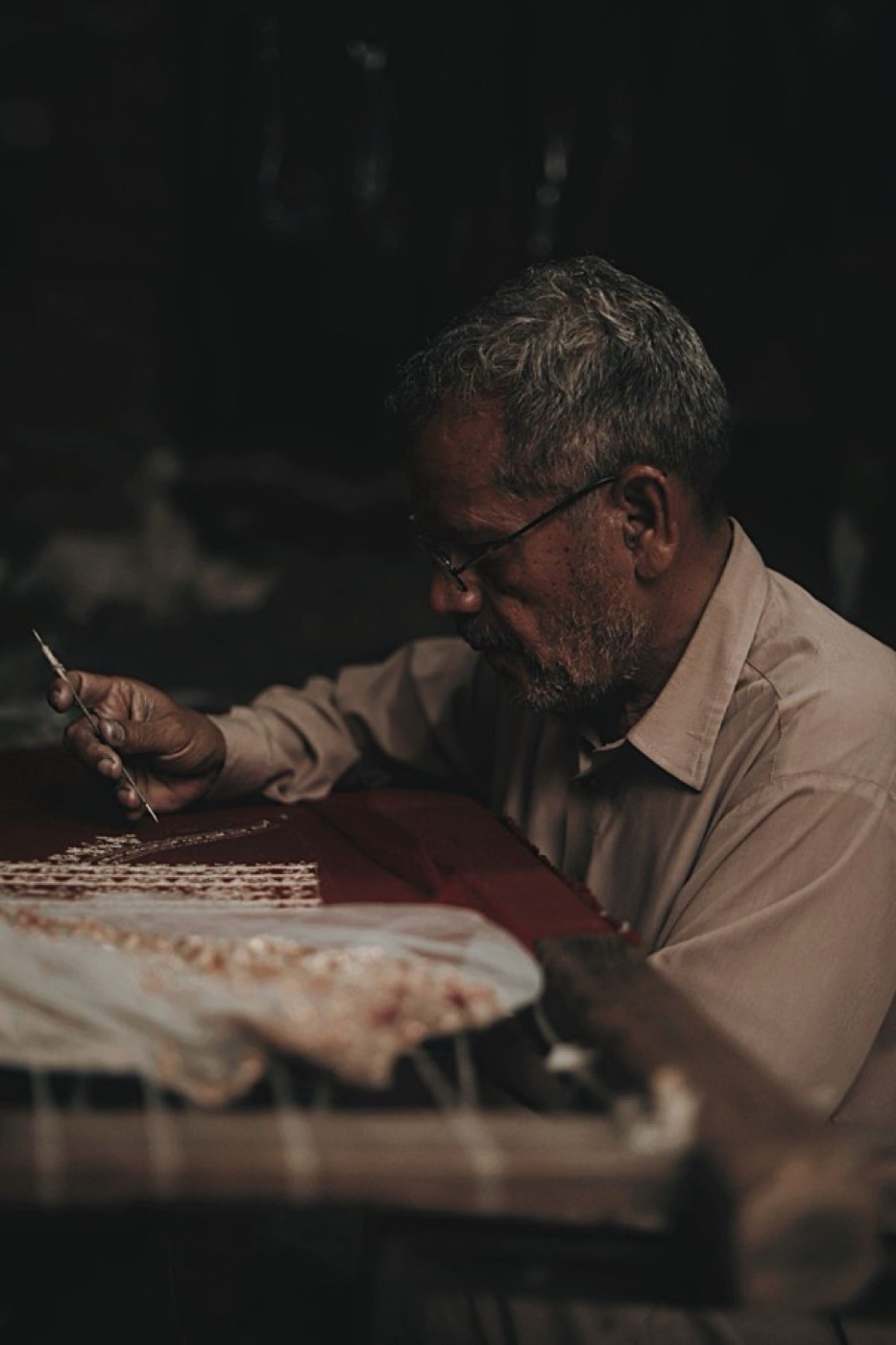 An artisan holding a fine aari hook needle above stretched fabric in a dim workshop